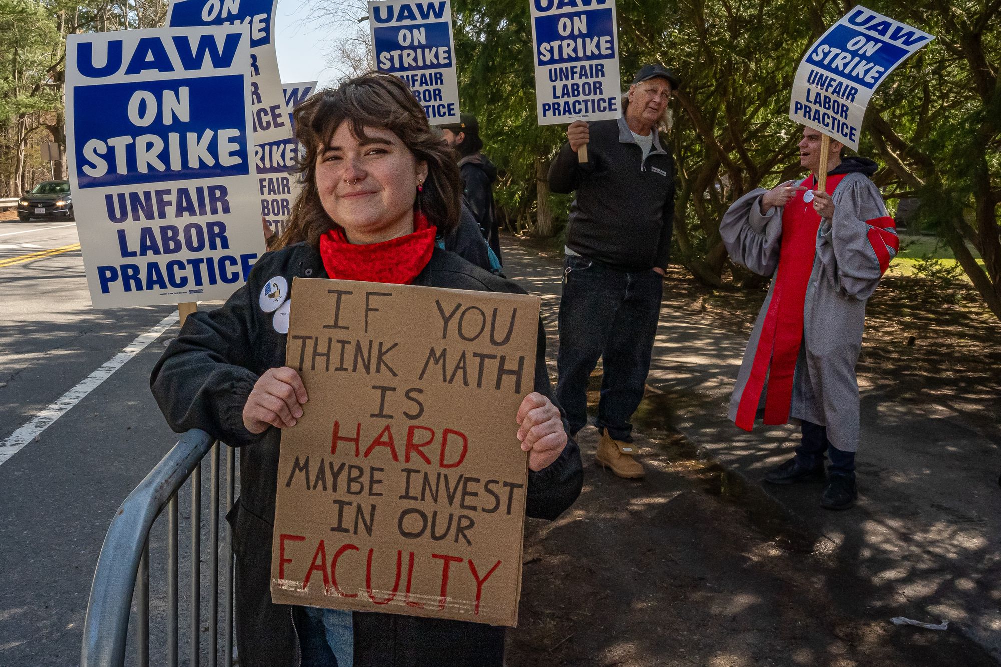 A young person holds a sign on a sidewalk that reads "If you think math is hard, maybe invest in our faculty." Other strikin