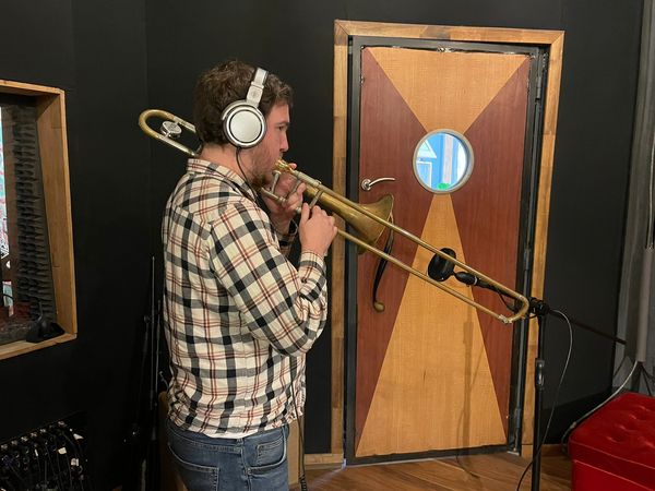 A male-presenting person plays a trombone wearing a plaid shirt, jeans, and headphones